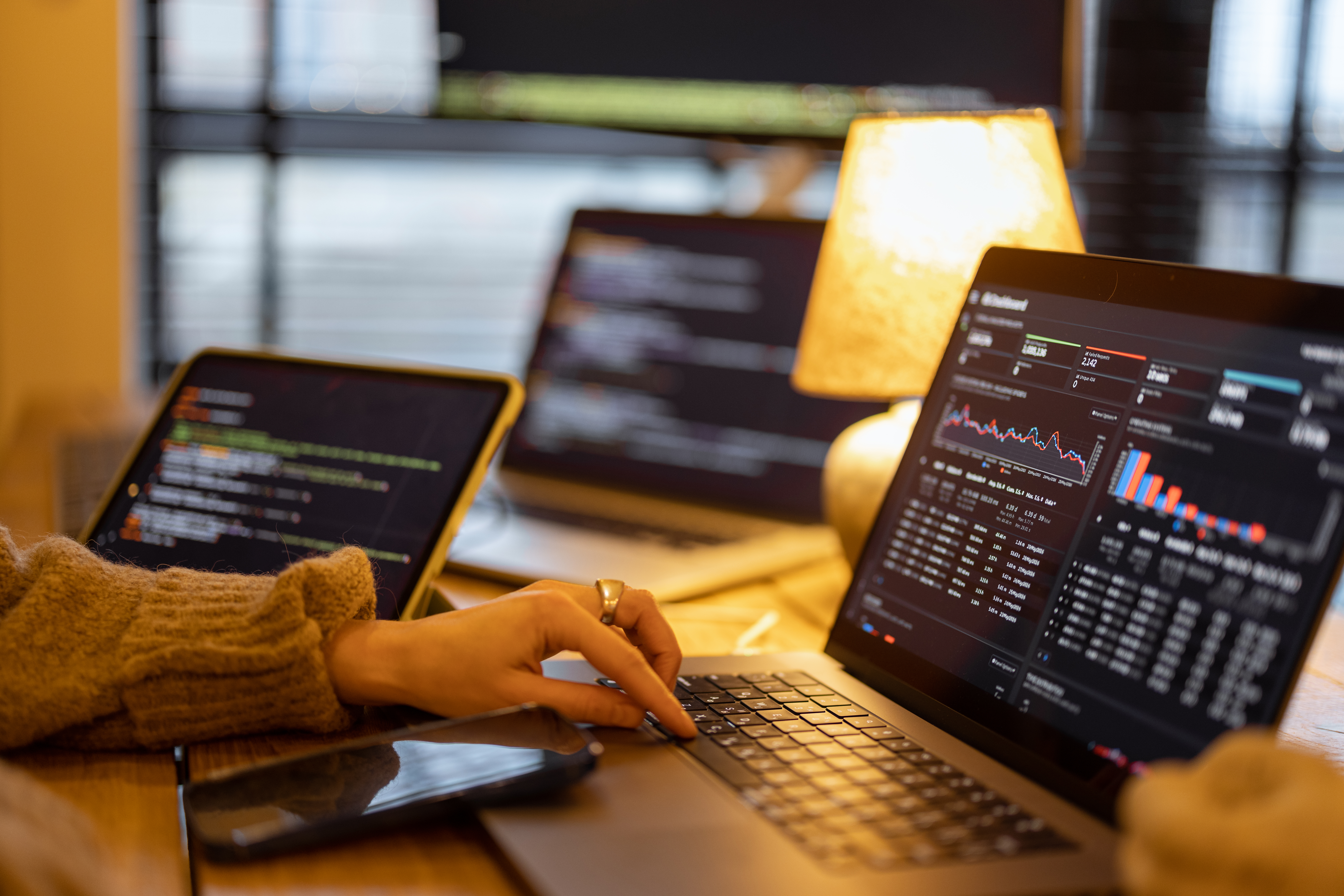 A person using multiple devices for programming or data analysis, showcasing a focused workspace illuminated by a lamp, with visual coding and statistical data displayed across the screens.